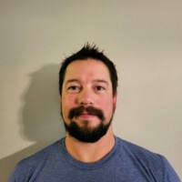 A man with short dark hair, a beard, and a mustache is wearing a blue T-shirt. He stands against a plain light-colored wall, smiling slightly at the camera.