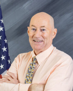 A bald man in a peach shirt and patterned tie is smiling with arms crossed. He stands in front of a backdrop featuring an American flag and a gray, textured background.