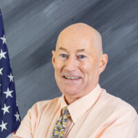 A bald man in a peach shirt and patterned tie is smiling with arms crossed. He stands in front of a backdrop featuring an American flag and a gray, textured background.