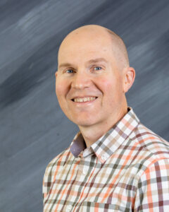 Reed Heidelberger, with a warm smile and a shaved head, stands confidently in a plaid shirt against a gray, textured background.