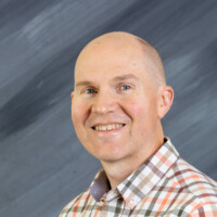 Reed Heidelberger, with a warm smile and a shaved head, stands confidently in a plaid shirt against a gray, textured background.