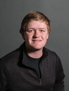 Kreg Anderson, with short, light hair, smiles slightly at the camera. They are dressed in a dark zip-up jacket over a black shirt. The background is a plain, dark gray.