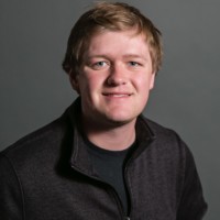 Kreg Anderson, with short, light hair, smiles slightly at the camera. They are dressed in a dark zip-up jacket over a black shirt. The background is a plain, dark gray.