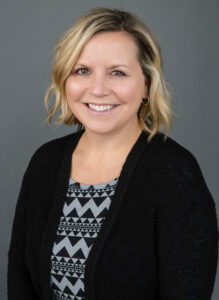 Michele Gelo, a smiling woman with shoulder-length blonde hair, wears a black cardigan over a gray patterned shirt as she poses gracefully against a plain gray background.