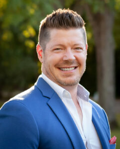 A smiling man with styled brown hair and a beard, wearing a blue suit jacket over a white shirt. He stands outdoors with a blurred background of trees and sunlight.