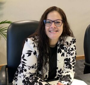 A person with long, dark hair and glasses is seated at a table. They are smiling and wearing a white blazer with a black floral pattern. The background is an office setting with a plant partly visible.