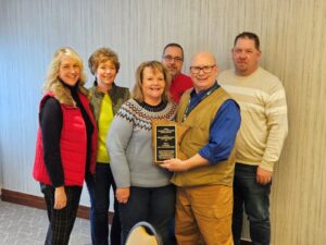 A group of six people smiling indoors, celebrating the Department of the Year award. One person proudly holds a plaque, and they all stand close together in winter clothing. The light-colored walls suggest a Building Department event, possibly AMBO 2023.