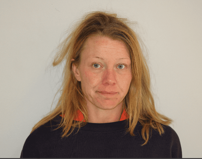 With tousled blonde hair and a neutral expression, Jasmin stands against a plain white background, wearing a dark sweater over a red top. She looks directly at the camera, her light skin softly illuminated.
